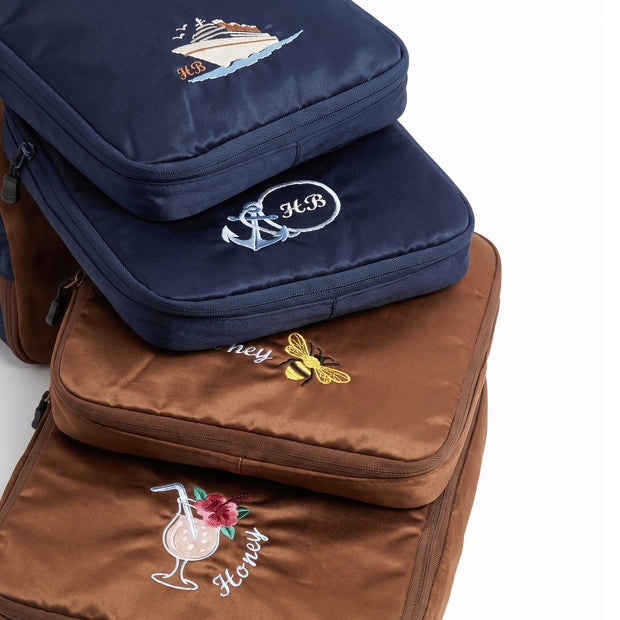 A stack of four caveri compression packing cubes in navy blue and brown, each featuring a different embroidered illustration and initials