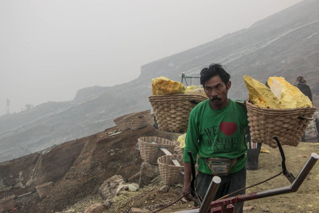 A miner carrying heavy sulphur blocks in baskets on his shoulders at Kawah Ijen, surrounded by yellow sulphur deposits